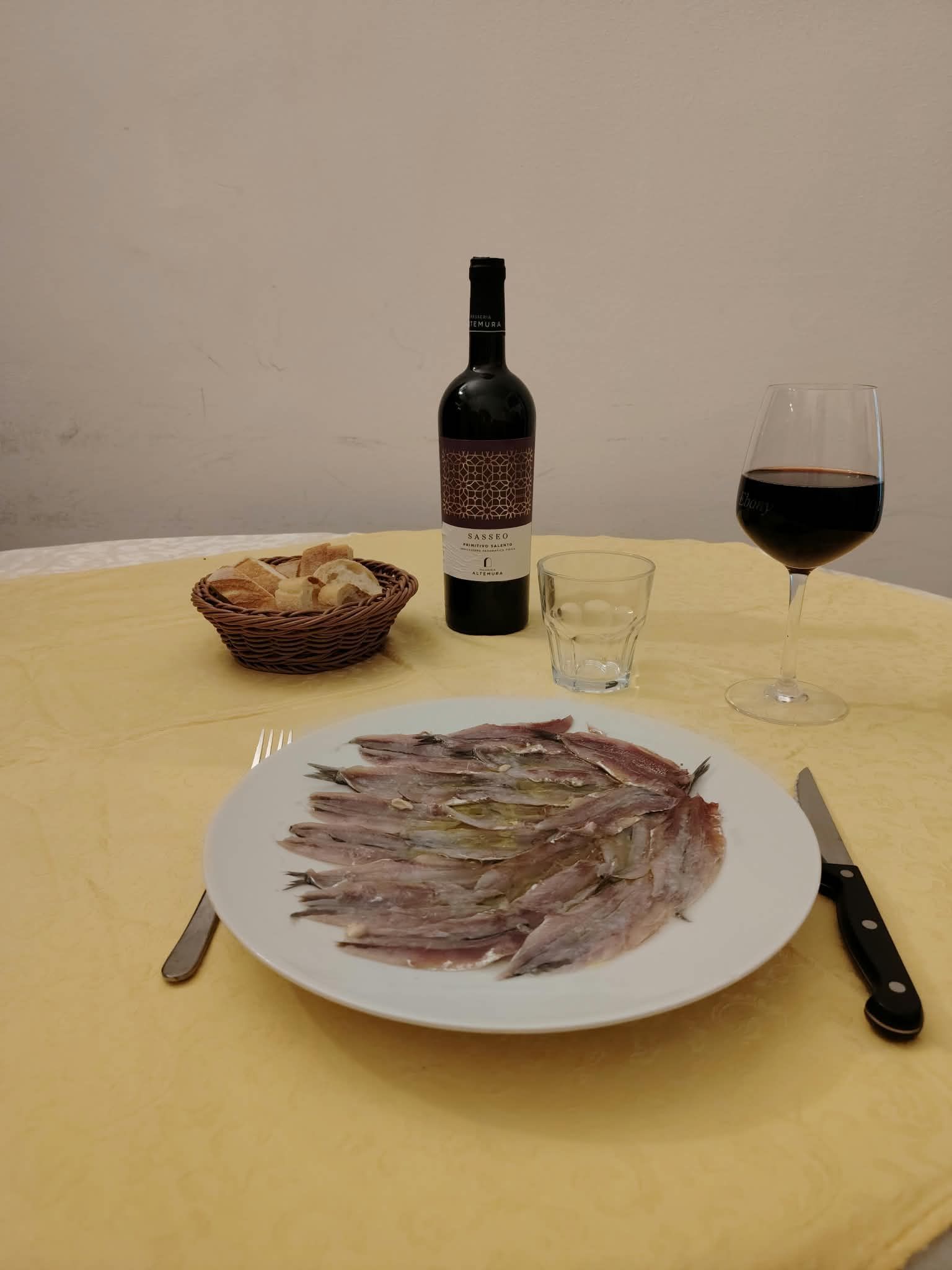Tavola apparecchiata con un pesce impiattato, un calice da vino, una bottiglia e un cestino del pane su una tovaglia gialla