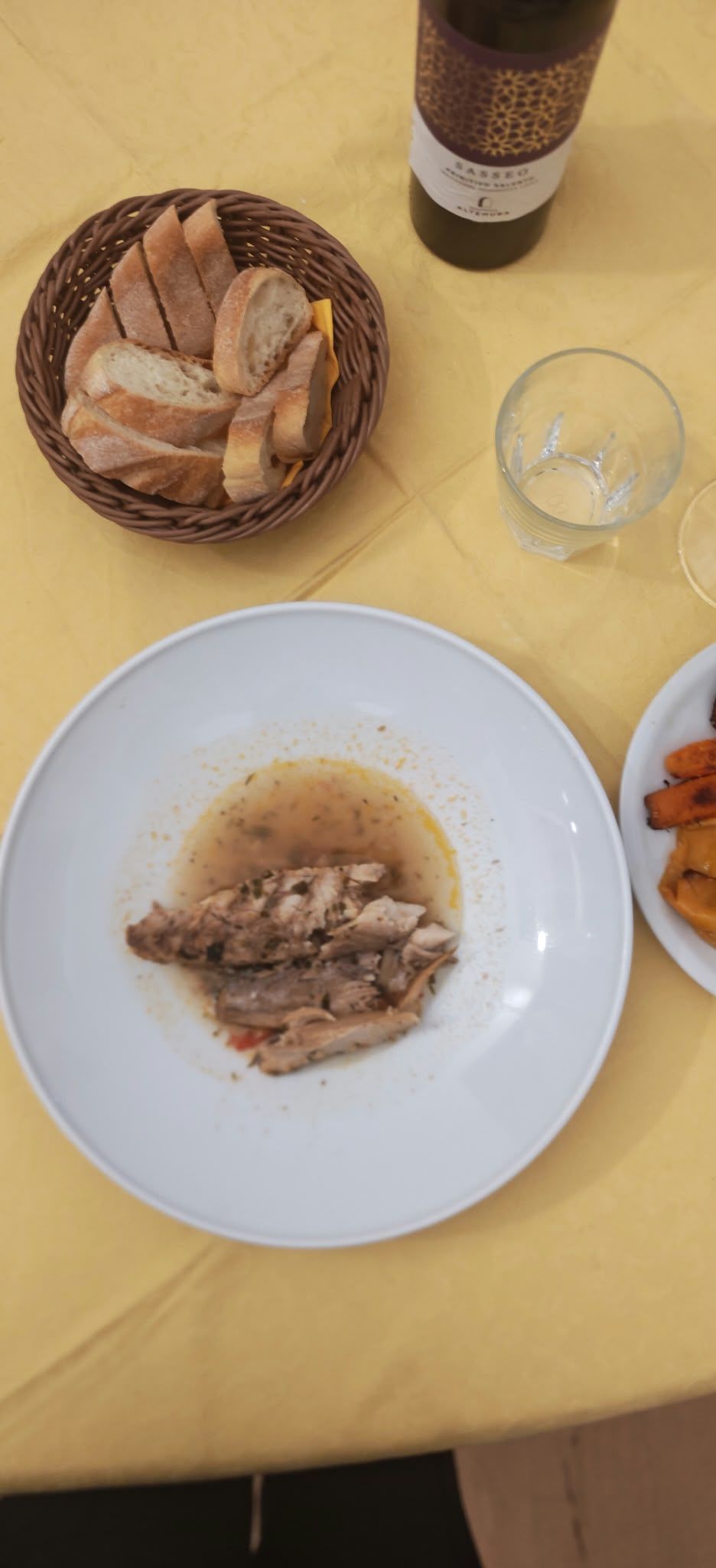 Tavolo con un piatto di pesce impiattato, cestino del pane, bottiglia di vino, bicchiere vuoto e patatine fritte su una tovaglia gialla