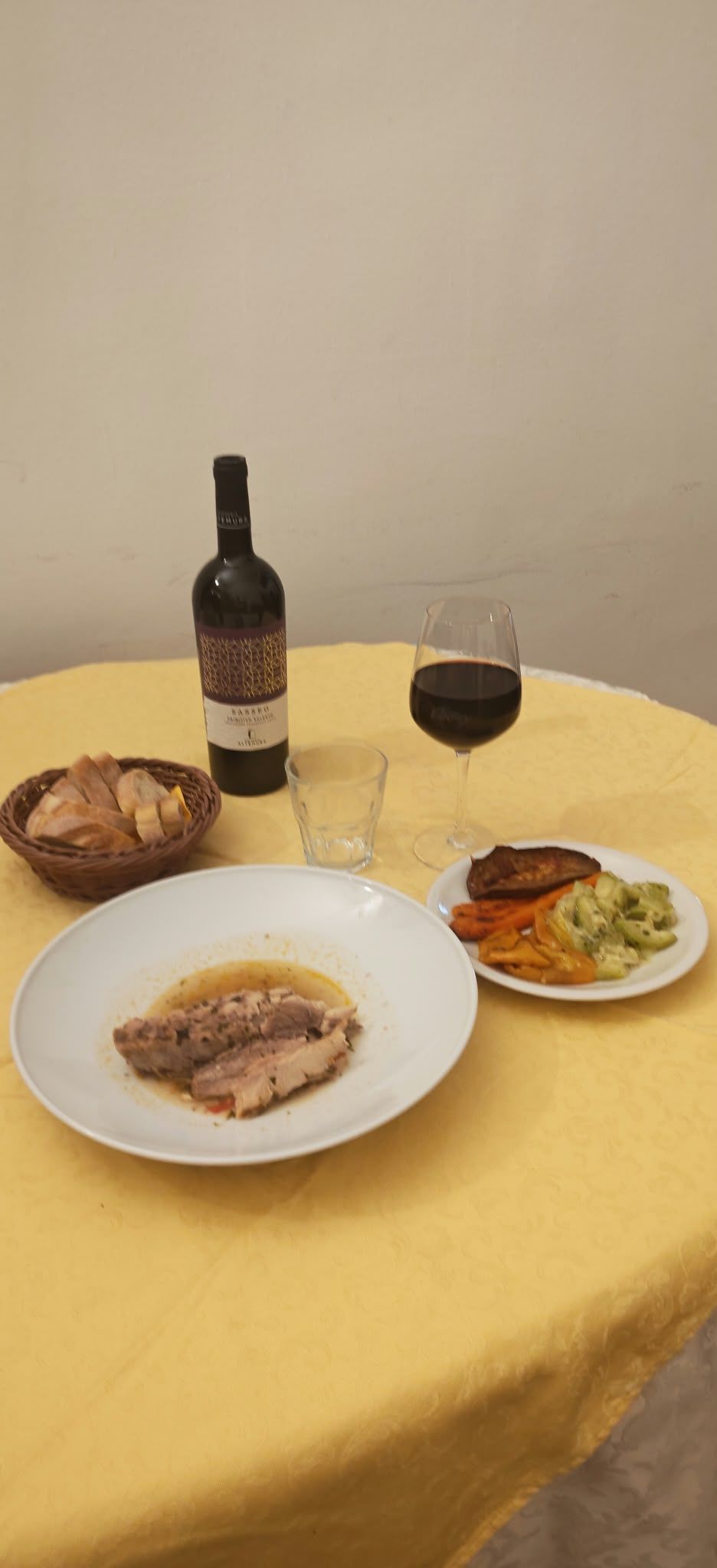 Tavola apparecchiata per un pasto con vino, pane, pasta e un piatto di carne su una tovaglia gialla.