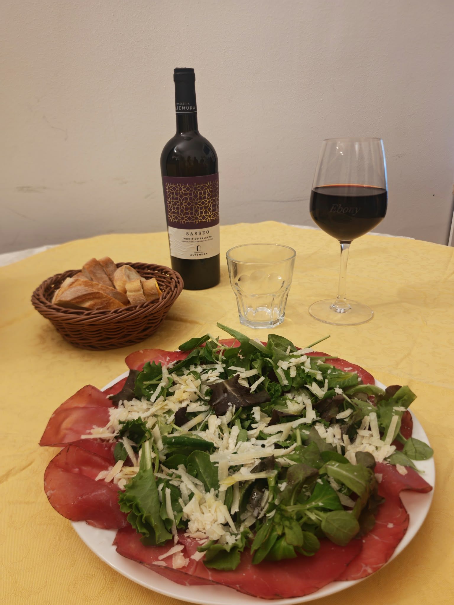 Tavola apparecchiata con insalata, bottiglia di vino, bicchiere di vino rosso, cestino del pane e un bicchiere vuoto