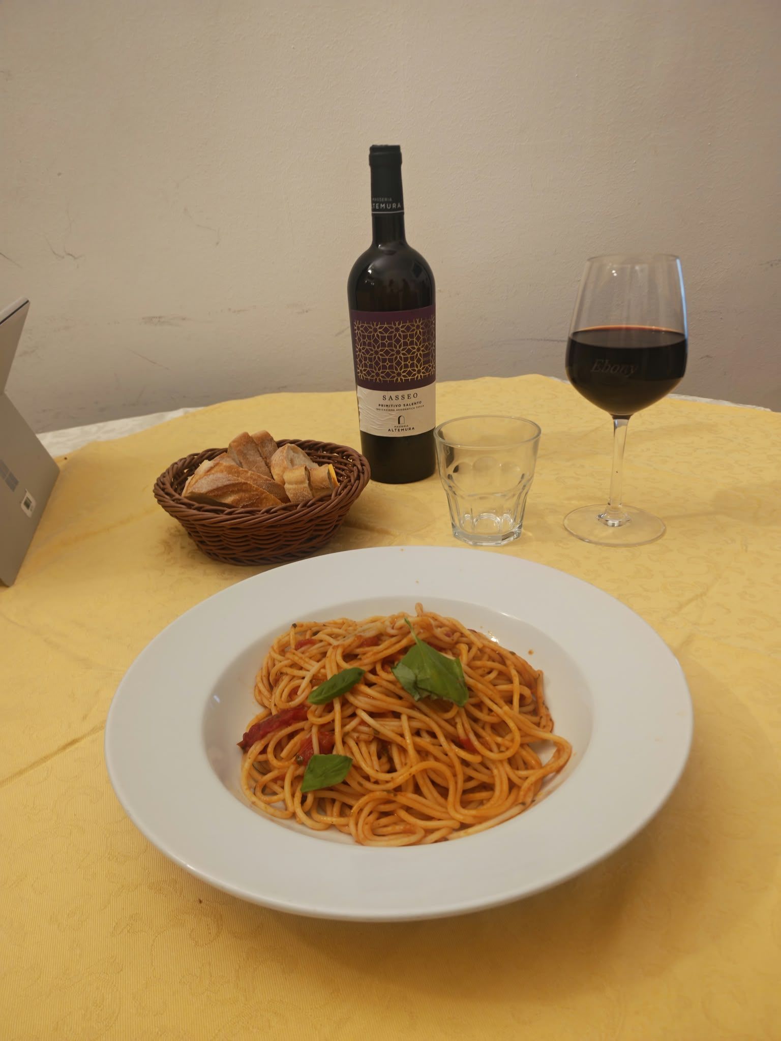 Piatto di spaghetti con basilico su un tavolo, accanto a una bottiglia di vino, un calice, un bicchiere d’acqua e un cestino del pane