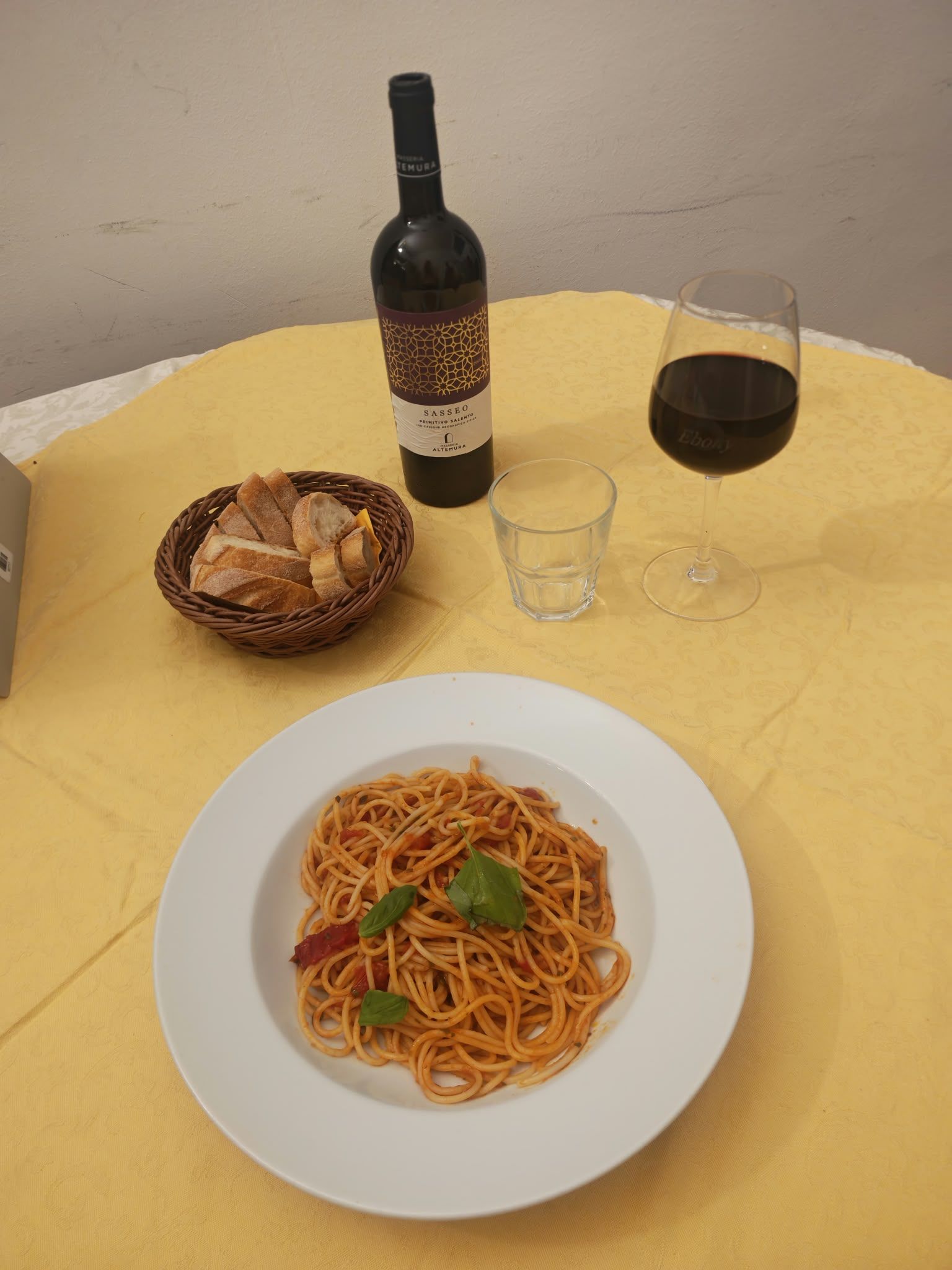 Pasta in una ciotola bianca su un tavolo giallo, con una bottiglia di vino, un bicchiere e un cestino del pane