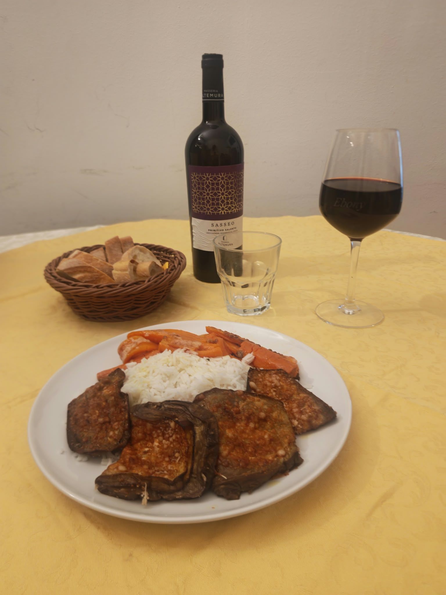 Una bottiglia di vino, un bicchiere e un cestino accanto a un piatto di carne alla griglia condita con salsa, su un tavolo.