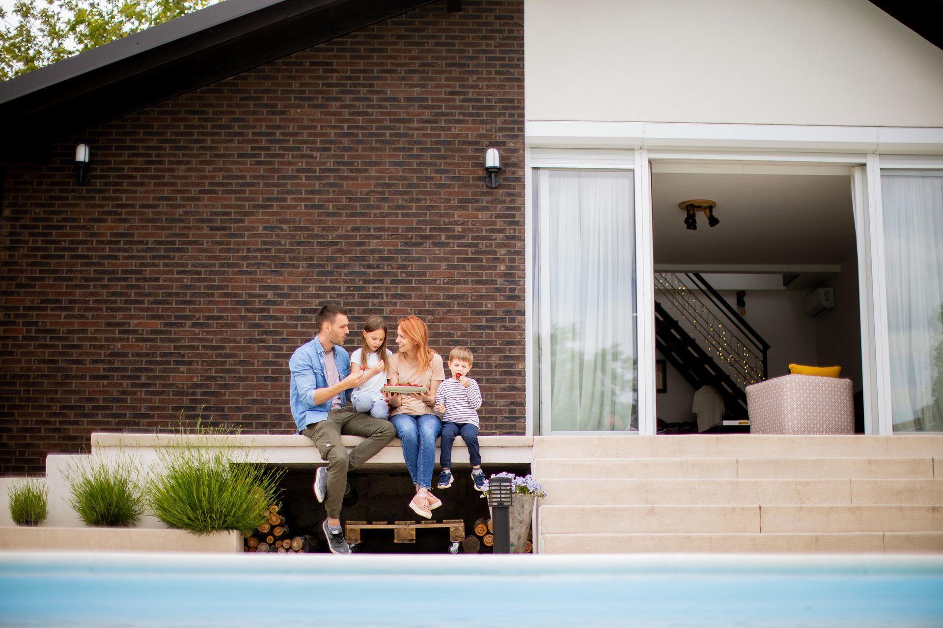 A family is sitting on the porch of their house next to a swimming pool.