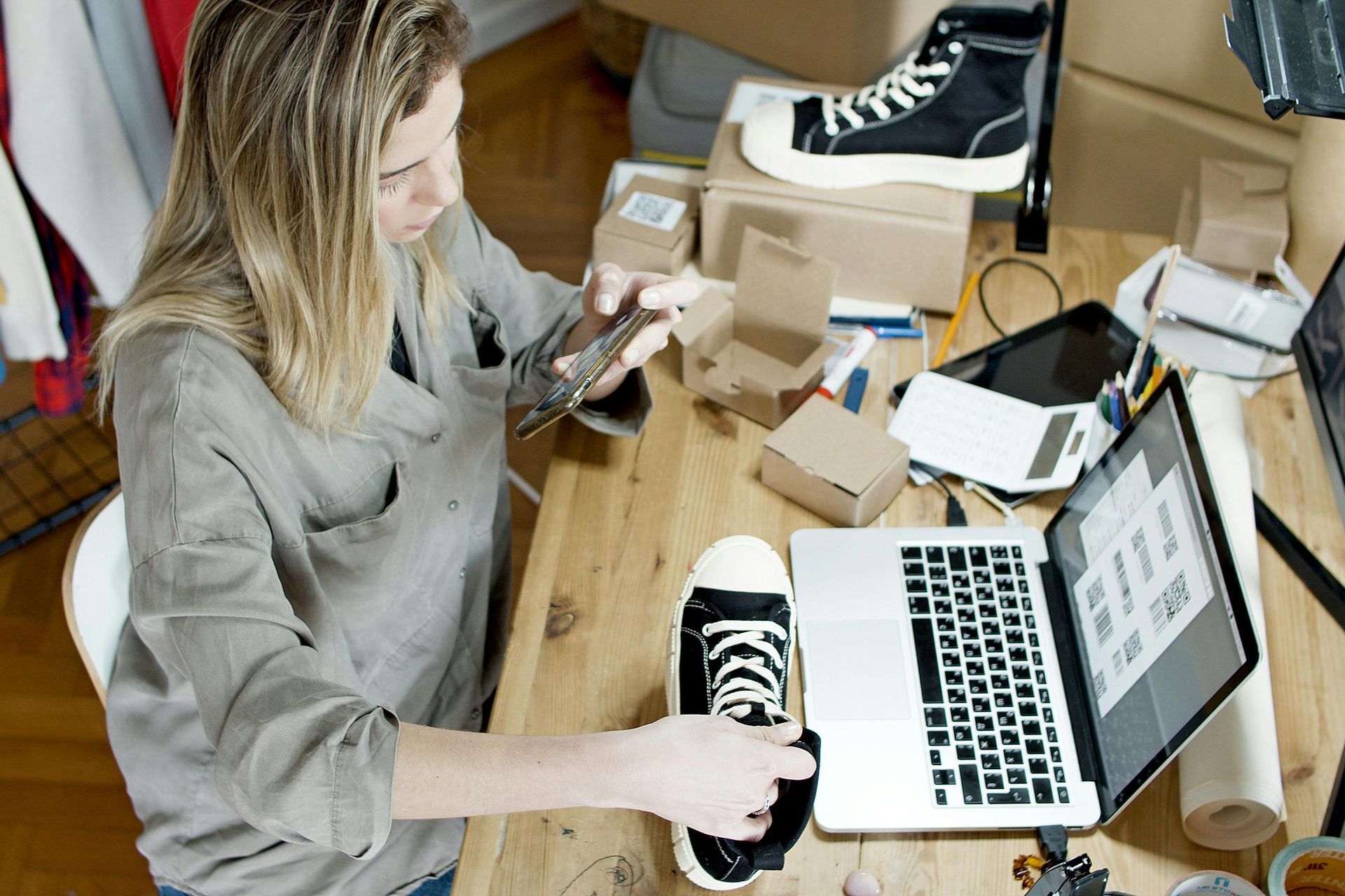 Woman taking photo of sneaker while working at a desk with laptop, boxes, and other shoes.