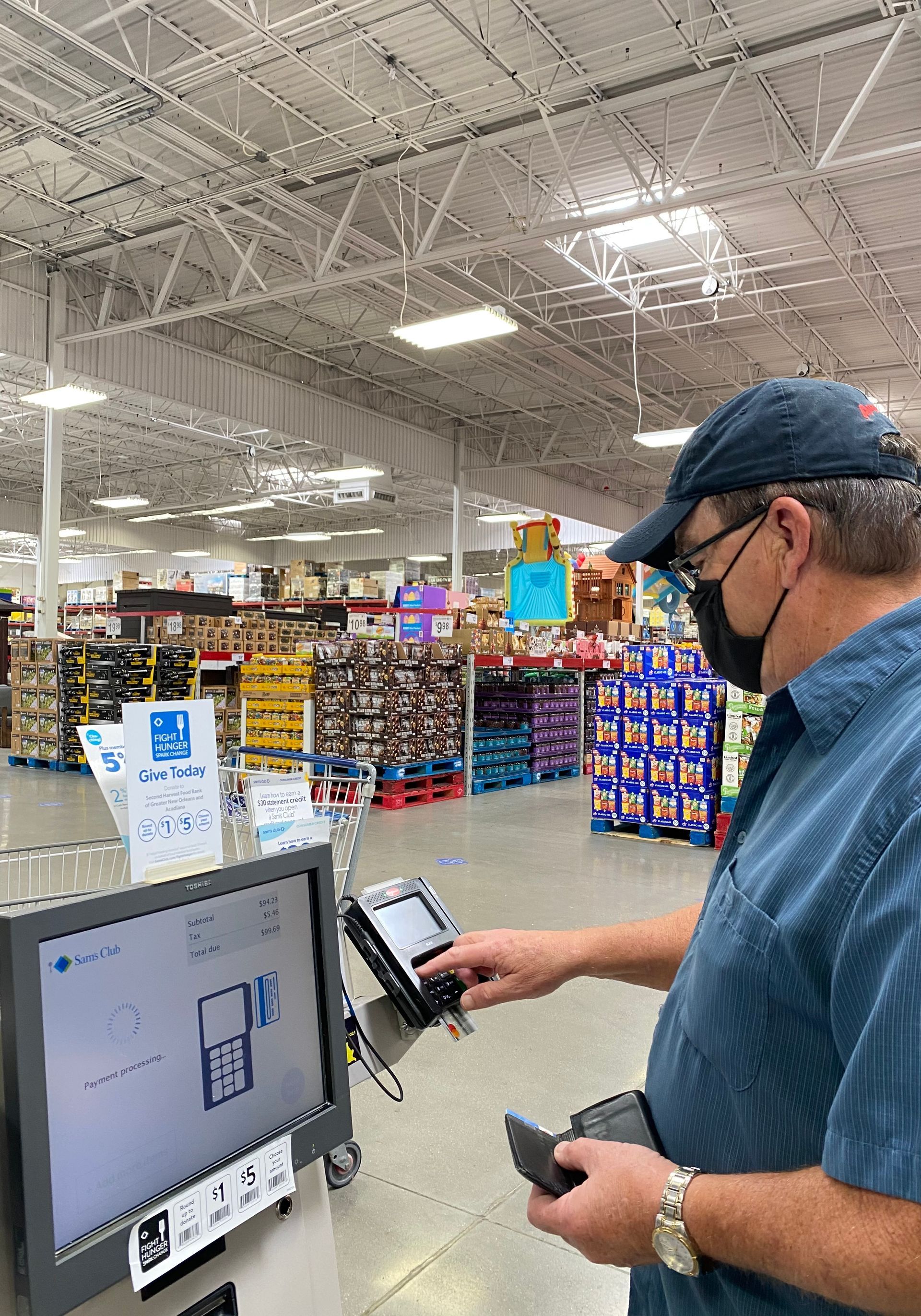 Man in mask using a self-checkout kiosk at a warehouse store, touching the payment screen.