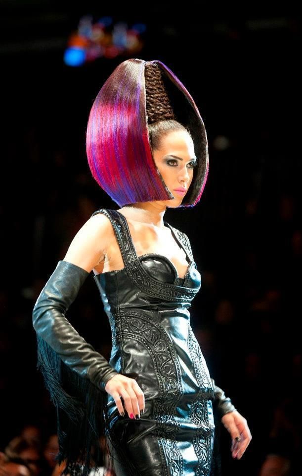 Fashion model in a shiny black outfit and long gloves, wearing a sculptural pink-and-purple hairstyle on a dark runway