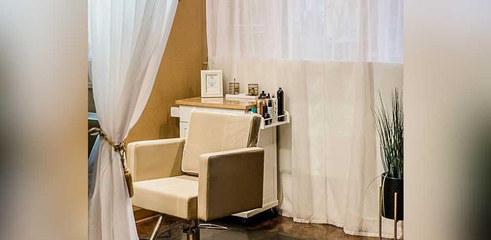 Empty beige salon chair beside a curtained window, with styling tools on a small counter