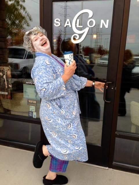 Woman in a blue patterned robe laughs outside a salon door, holding a drink and a small bag.