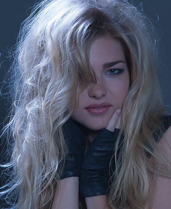 Portrait of a blonde person in black gloves, gazing at the camera against a dark blue background