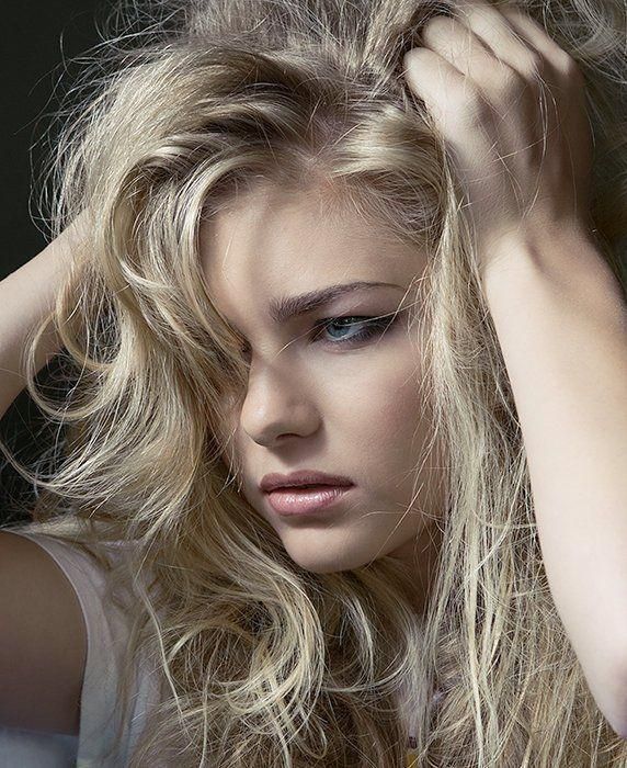 Close-up of a blonde person with tousled hair, resting a hand on their head and looking downward.