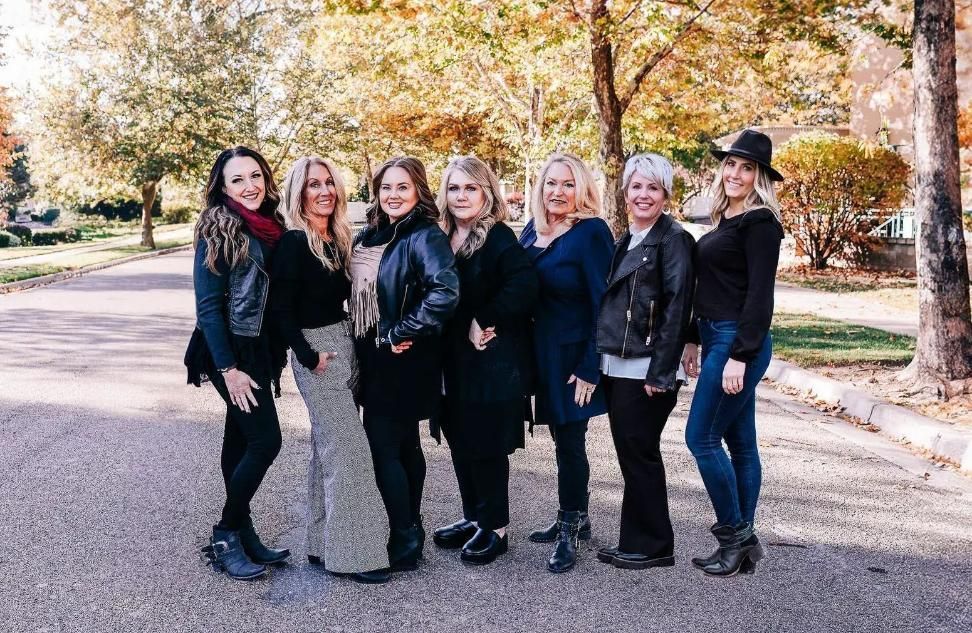 Seven people in black outfits posing outdoors on a tree-lined road in autumn.