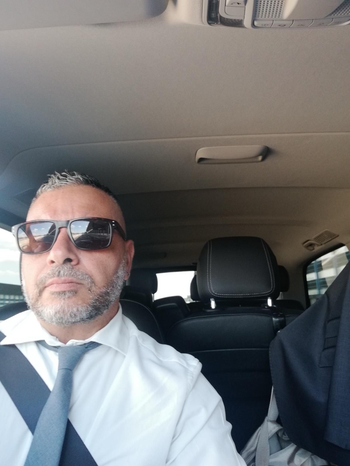 Un uomo con occhiali da sole e camicia bianca è seduto in macchina, con un altro passeggero parzialmente visibile.