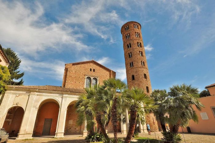 Edificio storico in pietra con ingresso ad arco e alta torre in mattoni sotto un cielo azzurro con palme