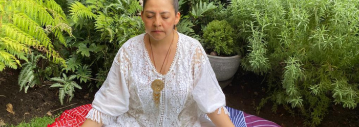 Una mujer medita al aire libre, rodeada de vegetación. Viste de blanco, con los ojos cerrados y un colgante.