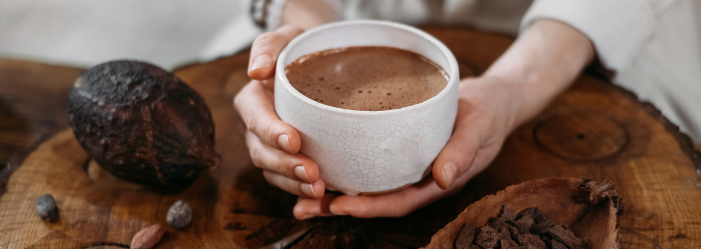 Manos sosteniendo una taza de chocolate blanco, con granos de cacao y vainas sobre una superficie de madera.