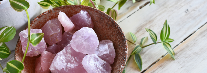 Cristales de cuarzo rosa en un recipiente marrón rodeado de plantas de hojas verdes y blancas sobre una superficie de madera blanca.