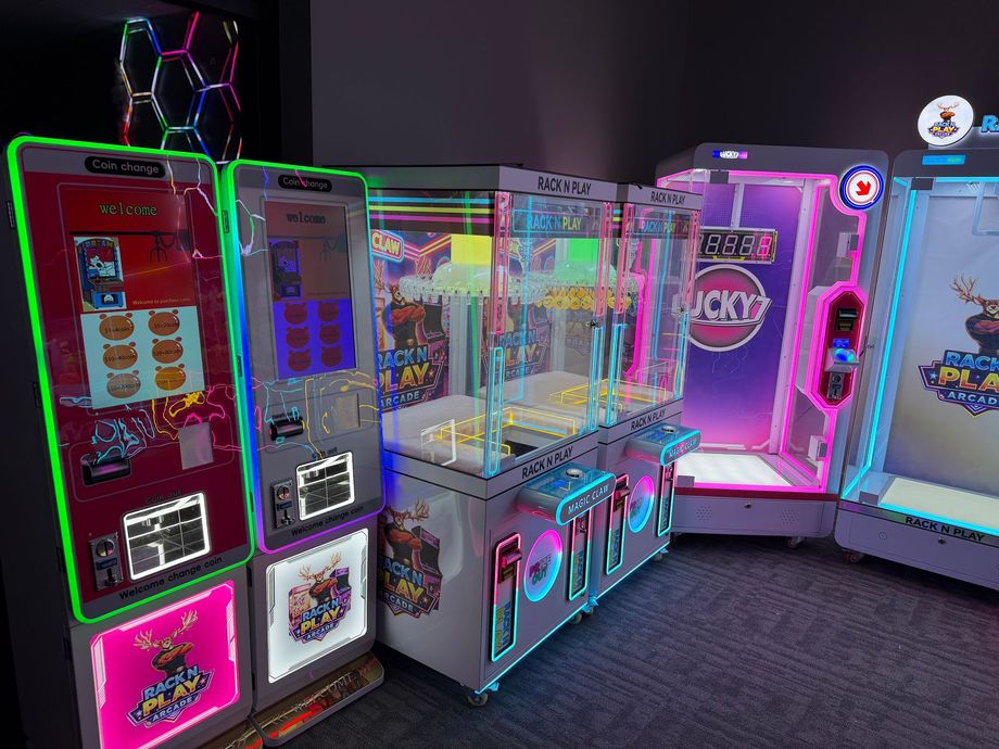 Neon-lit arcade claw machines in bright pink, blue, and silver lined up in a game room.
