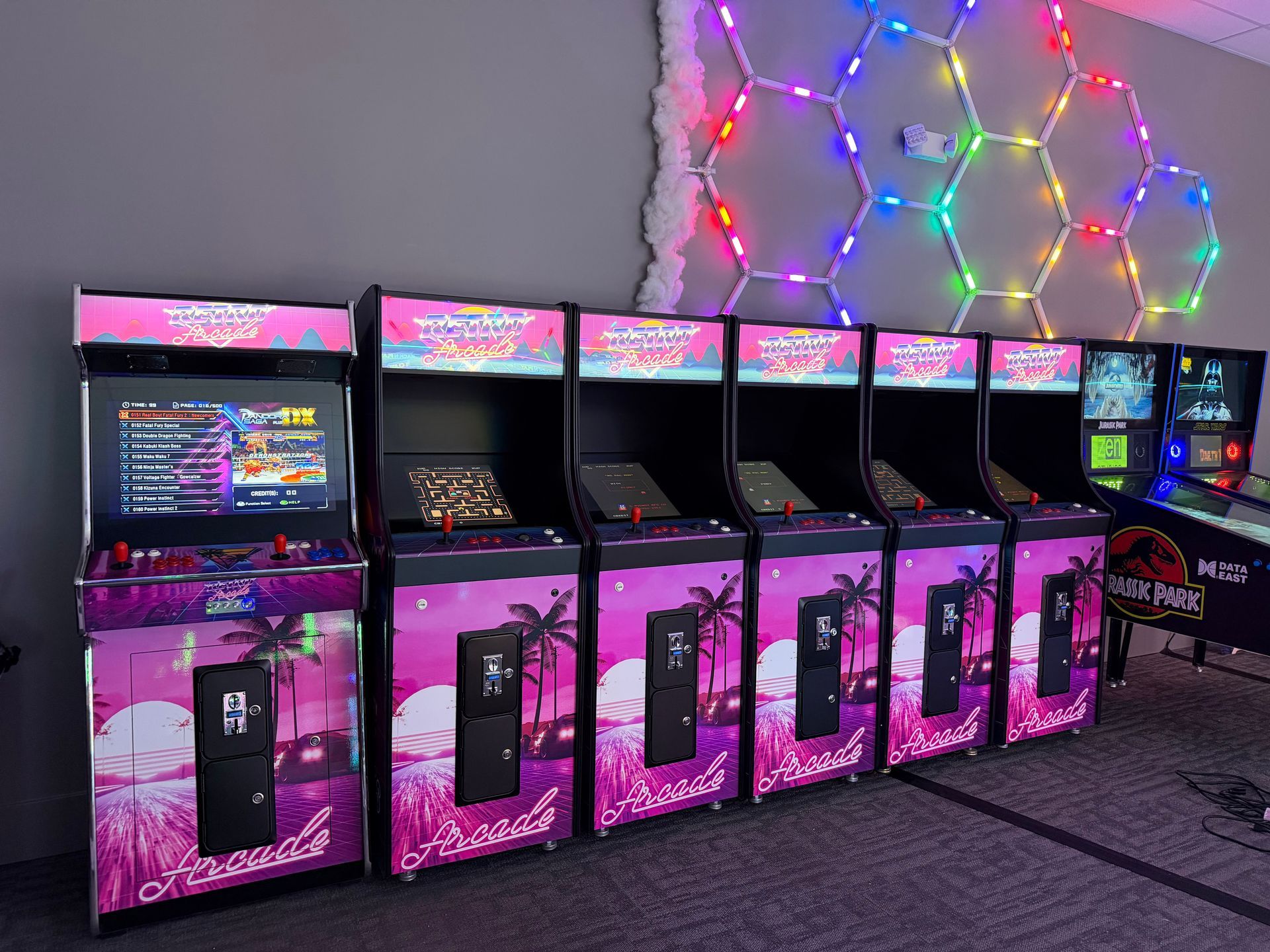 Row of colorful arcade cabinets with pink-lit fronts and neon wall lights in a dark room