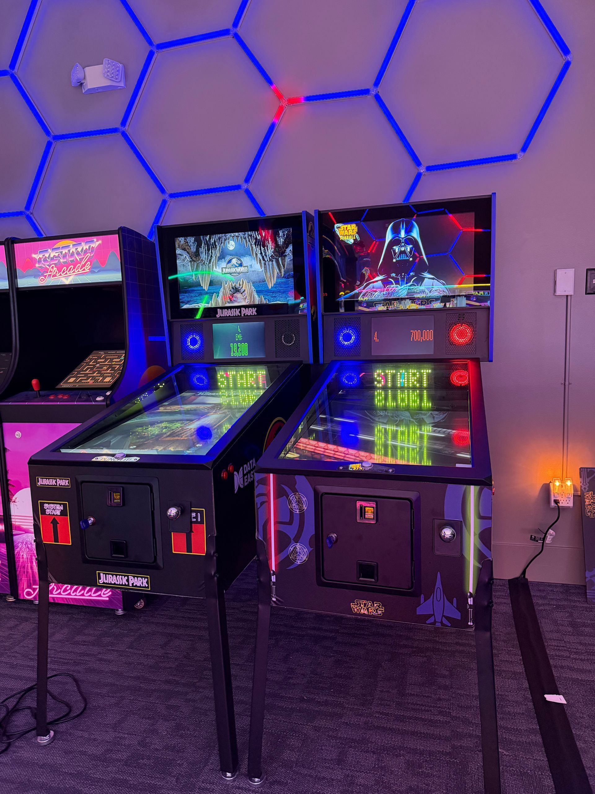 Two illuminated arcade cabinets glowing blue and pink in a dim room with hexagonal wall lights.