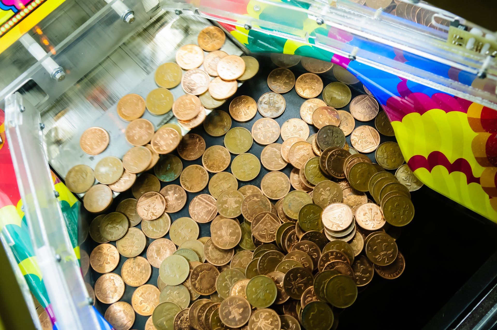 Coin pusher arcade game tray filled with scattered pennies and coins under colorful plastic panels