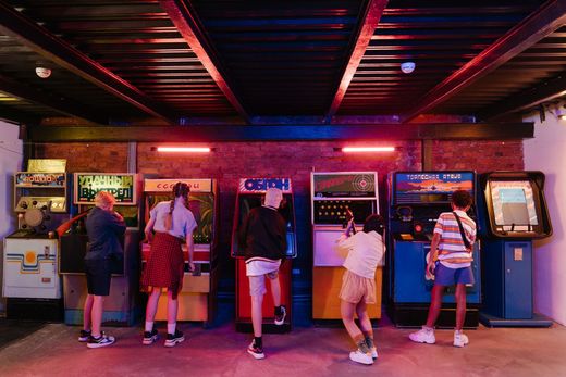 People playing arcade games in a neon-lit arcade with brick walls and colorful cabinets