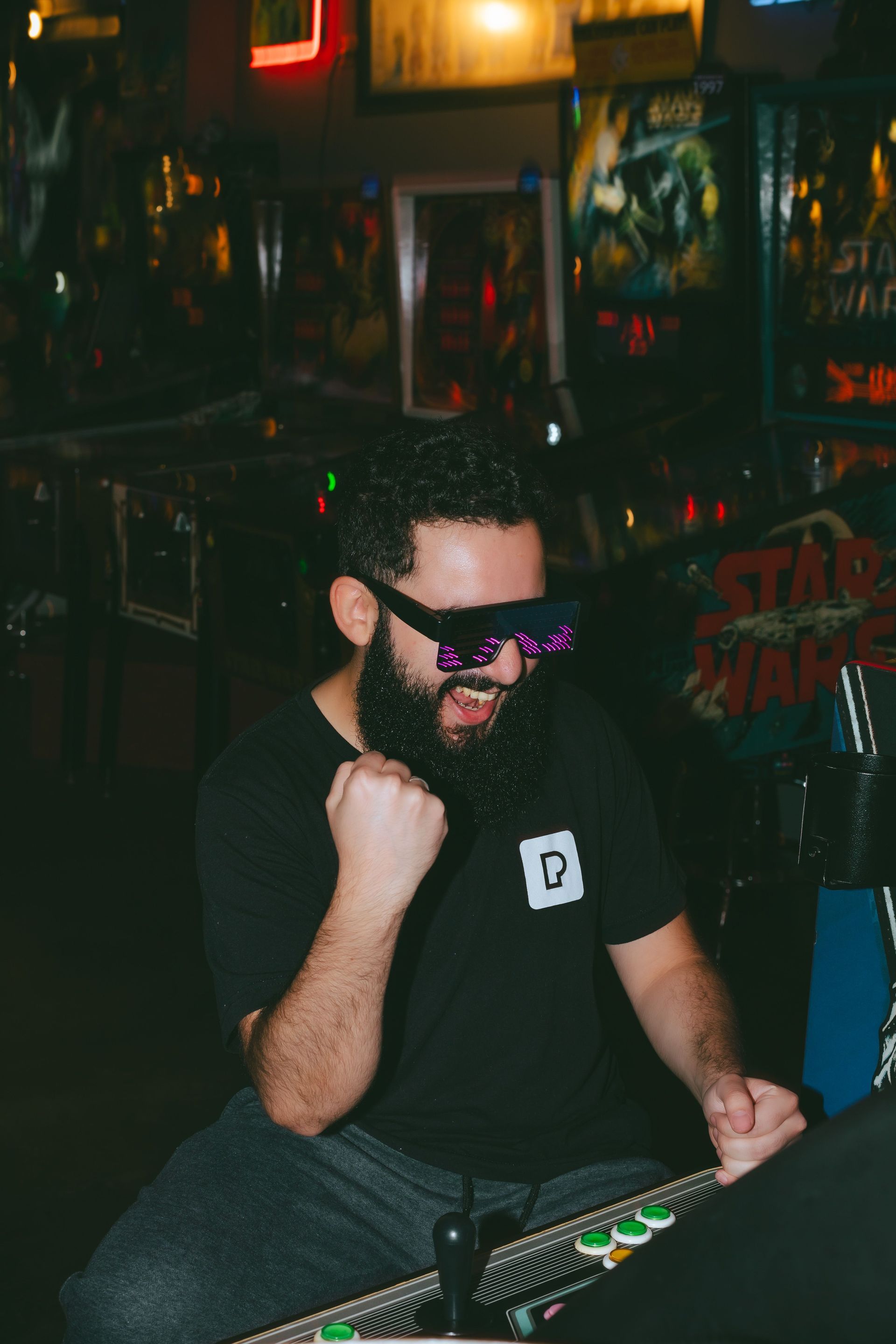 Man with sunglasses and beard seated at an arcade machine in a dimly lit bar.