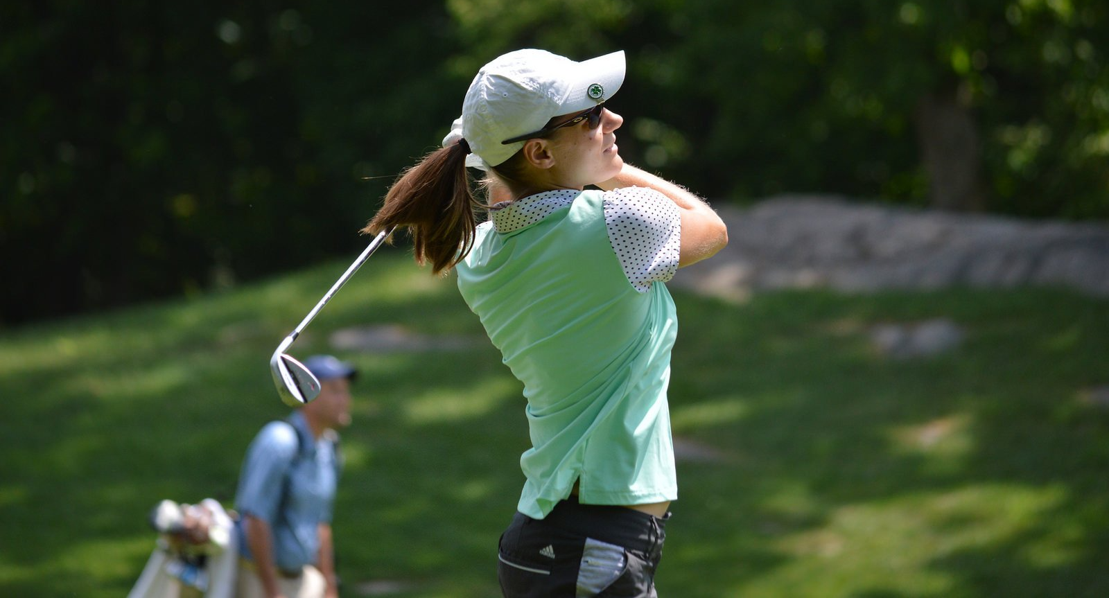 Woman in a light green shirt and white cap swings a golf club on a sunny course. A man watches in the background.