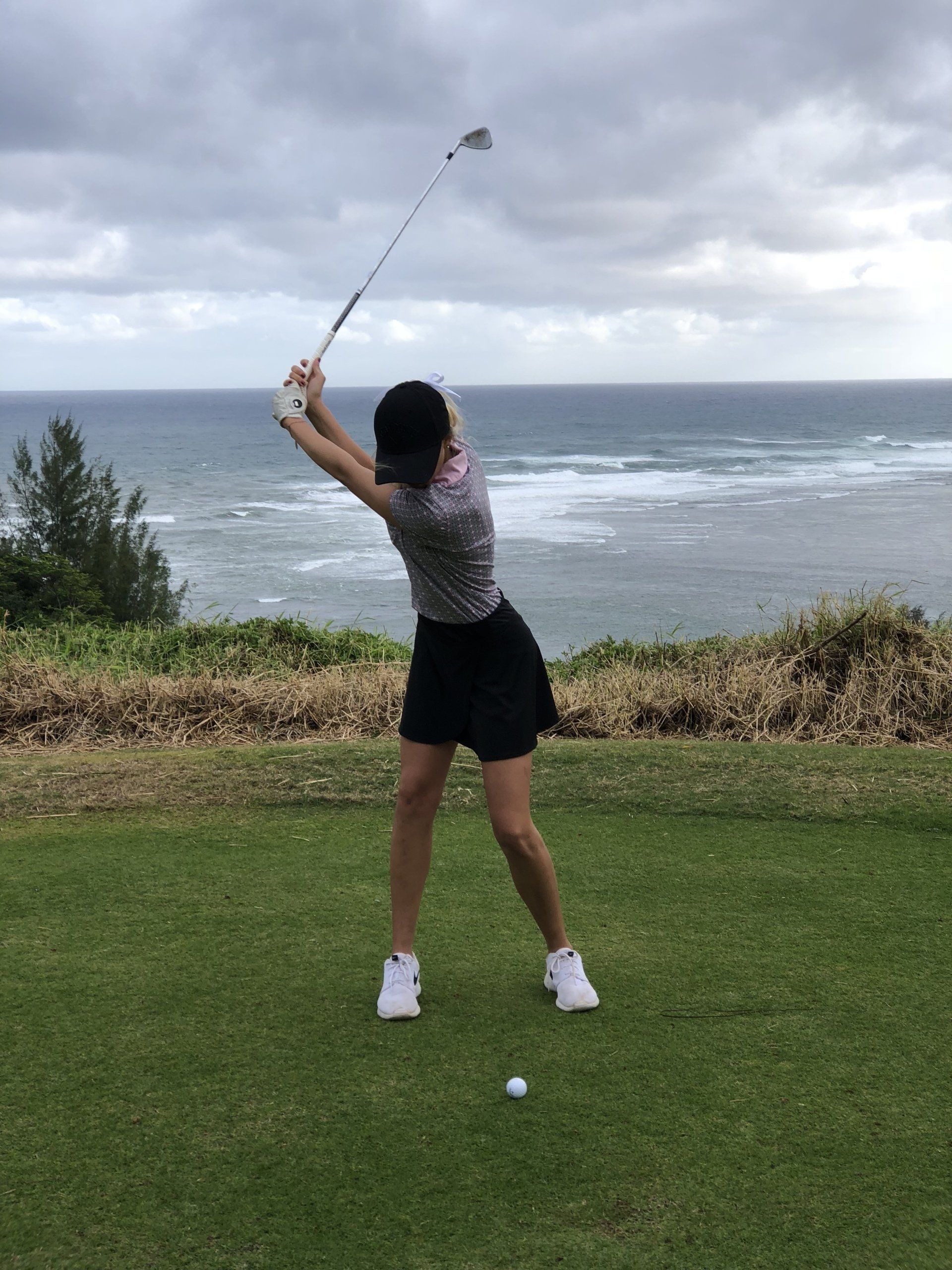 Woman in golf attire swinging a club on a green, ocean in background. 