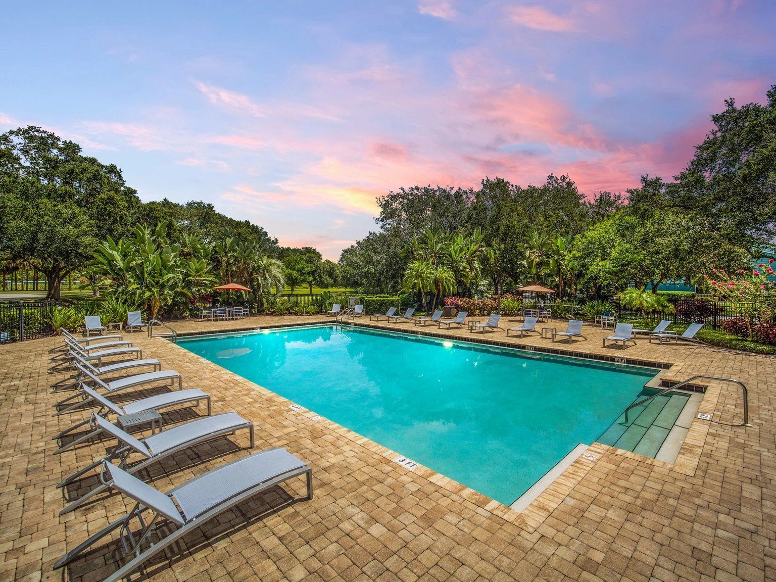 Swimming pool surrounded by lounge chairs, trees, and brick patio under a colorful sunset.