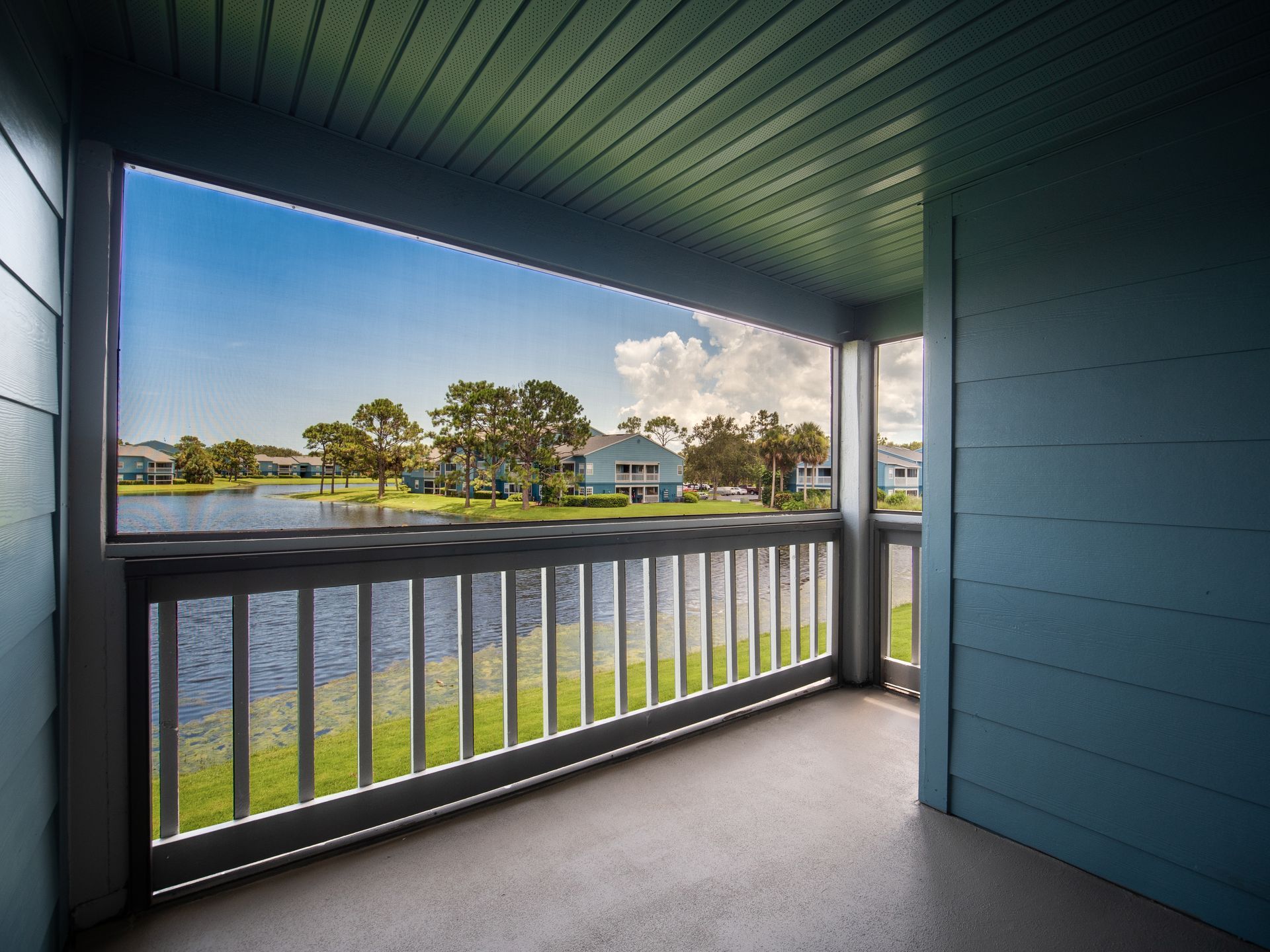 Balcony overlooking a lake and houses under a blue sky, railing and walls are light blue. Experience comfortable living in our Melbourne, FL apartments for rent—schedule your walkthrough today.
