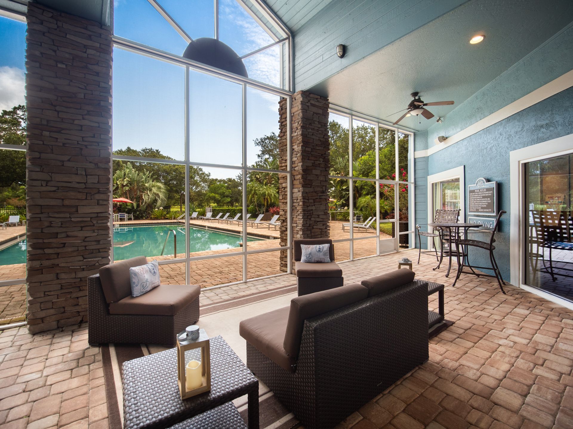 Clubhouse patio with pool view: Brown wicker furniture, large windows, brick floor, blue walls, ceiling fan, and pool outside. Come experience these thoughtfully designed Apartments in Melbourne, FL in person with a guided tour.