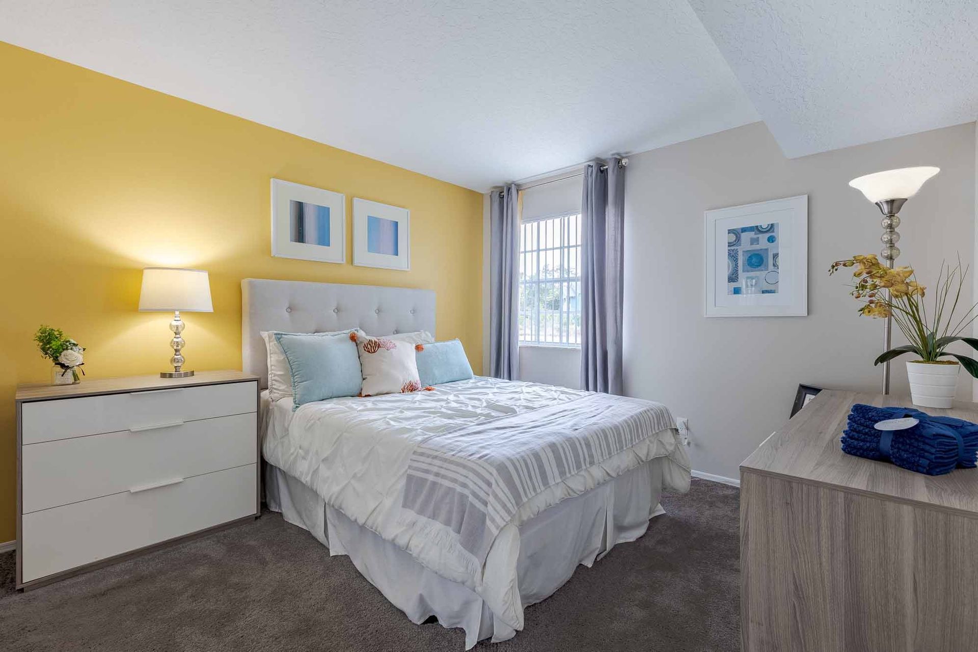 Bedroom with yellow accent wall, gray dresser, bed with white bedding, and two framed pictures. Take a closer look at these welcoming Apartments in Melbourne, FL by scheduling your tour today.