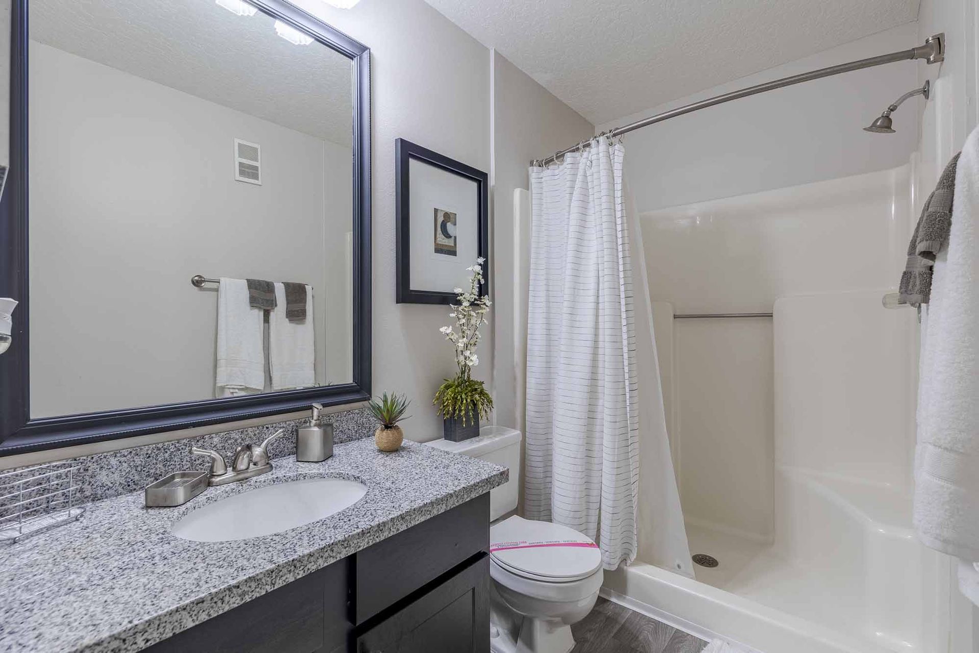 Bathroom with a granite countertop, large mirror, shower, and toilet. White, grey, and black color scheme. Tour our beautifully crafted Apts for rent in Melbourne, FL and find your perfect fit.