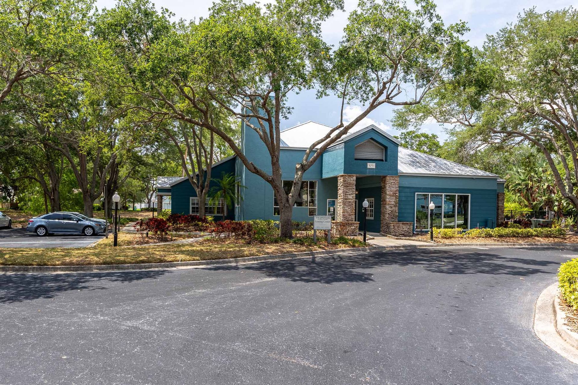 Exterior resident clubhouse view. Blue building with stone accents, surrounded by trees, parked cars, asphalt driveway. Discover the comfort of our Pet-friendly aparmtents in Melbourne, FL with an in-person tour.
