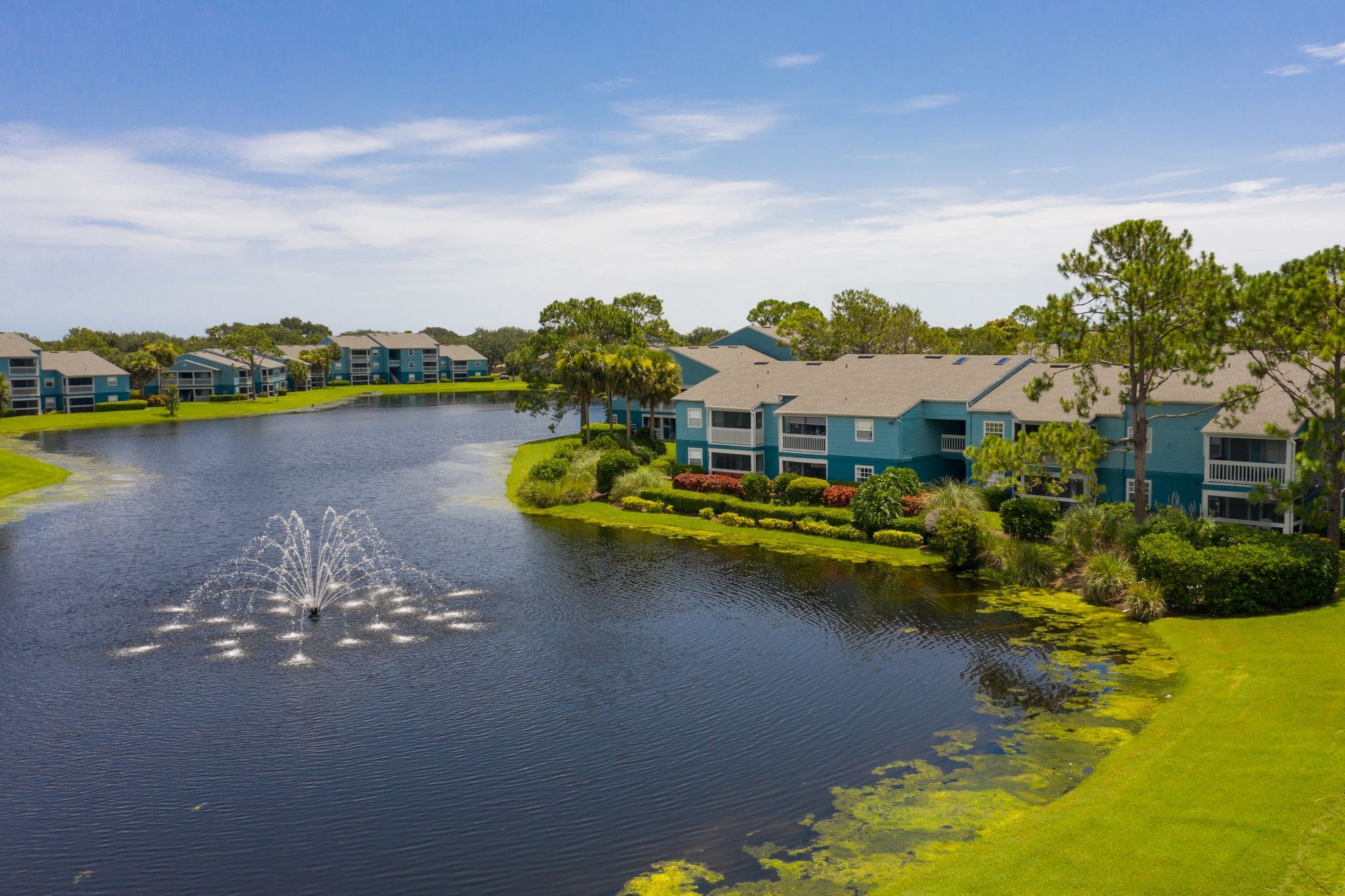 Aerial view of blue apartment buildings on a lake with a fountain and green landscaping. Visit Savannahs at James Landing and explore our welcoming Apts for rent in Melbourne, FL today.