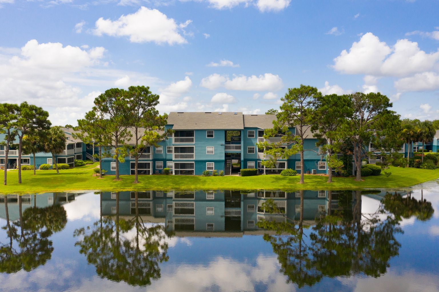Blue apartment building reflected in still water, surrounded by trees under a partly cloudy sky. Tour these beautifully maintained Apartments in Melbourne, FL and see what makes them special.