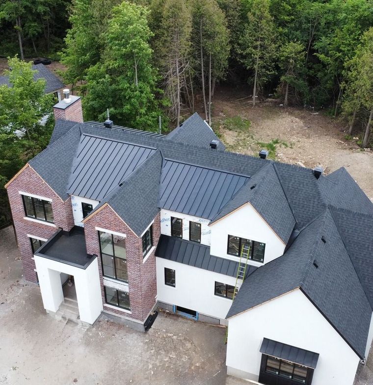 Aerial view of a large modern house with dark gray roofs, white and brick siding, beside a wooded area
