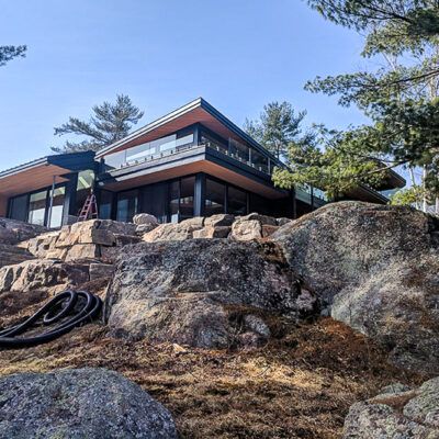 A modern, flat-roofed house built on a rocky, uneven landscape surrounded by pine trees under a clear blue sky.