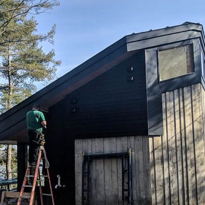 A person in a green shirt stands on a ladder working on the black siding of a modern home with wood-paneled walls.