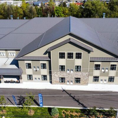 An aerial view of a multi-story tan commercial building with a grey metal roof and stone accents, fronted by a road.
