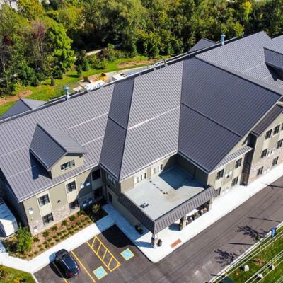 An aerial view of a multi-story building with a gray metal roof, attached parking lot, and surrounding green landscape.