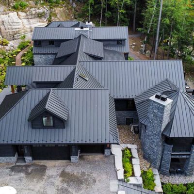 An aerial view of a large modern house with dark metal roofing and stone walls, situated in a wooded, rocky landscape.