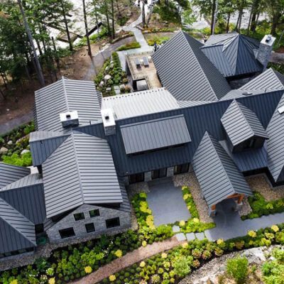 Aerial view of a large modern house with a dark grey metal roof, stone walls, and surrounding lush landscaping.
