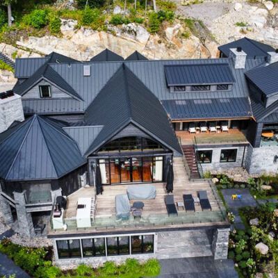 An aerial view of a luxury multi-level house with a dark metal roof, stone exterior, and a spacious wooden patio deck.