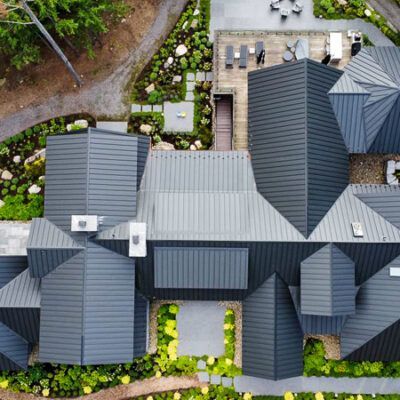 Aerial view of a large modern house with a dark metal roof, surrounded by landscaping, a patio, and a driveway.