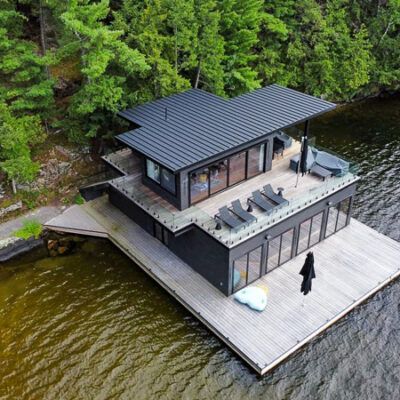A modern, black-sided house sits on a wooden dock over a lake, surrounded by lush green trees.