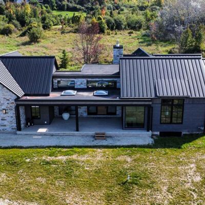 A modern house with a black metal roof and stone accents sits on a grassy hill surrounded by trees.