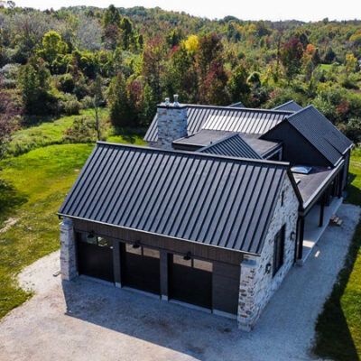 An aerial view of a modern stone and dark-clad house with a black metal roof, nestled in a lush, green countryside.