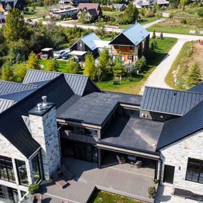 Aerial view of a contemporary home with stone walls and a black metal roof, featuring a patio and surrounding landscape.